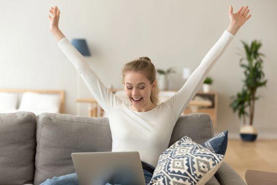 Frau-reisst-mit-Laptop-auf-dem-Schoss-vor-Freude-die-Arme-in-die-Luft